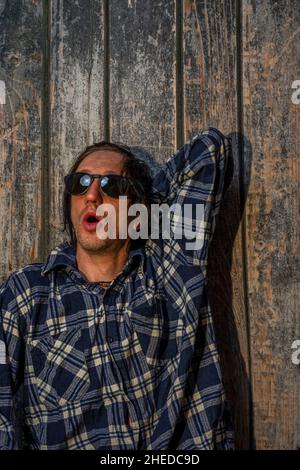 Uomo di capelli neri seduto vicino a una vecchia parete di legno con camicia in serata calda al tramonto Foto Stock