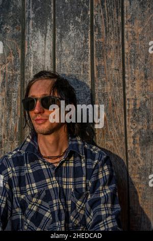 Uomo di capelli neri seduto vicino a una vecchia parete di legno con camicia in serata calda al tramonto Foto Stock