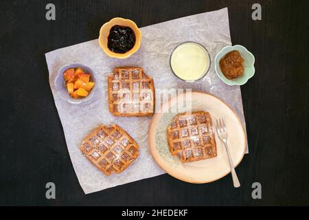 Vista dall'alto di vegan, cialde senza glutine con guarnizioni di marmellata e frutta e sfondo nero di zucchero in polvere. Sana e gustosa colazione fatta in casa. Foto Stock