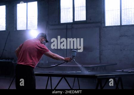 Vista frontale dell'uomo adulto in T-shirt rossa con cappuccio nero che trafila la recinzione attraverso il polivizor in grande officina spaziosa. Concetto di uomo che lavora con polivizor su sfondo bianco. Foto Stock