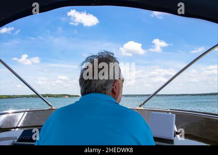 Vista ravvicinata posteriore dell'uomo dai capelli grigi in occhiali da sole che guida una barca sul lago con case e litorale all'orizzonte sotto il cielo nuvoloso blu Foto Stock