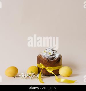 Una dolce pasquale festosa cosparso di zucchero in polvere e legato con un nastro di raso giallo e fiori bianchi e uova gialle dipinte su un backgroun beige Foto Stock