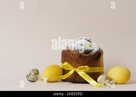 Una dolce pasquale festosa cosparso di zucchero in polvere e legato con un nastro giallo satinato e fiori bianchi e uova dipinte di giallo e quaglia su un beige Foto Stock