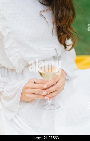 Un bicchiere di vino bianco o champagne nelle mani di una giovane donna in un abito bianco. Primo piano Foto Stock