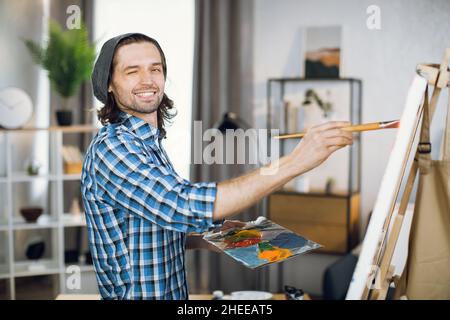 Felice artista maschile che vagliano e sorride sulla fotocamera mentre disegnano con pennello su tela. Bel ragazzo caucasico godendo di processo creativo in studio luminoso. Foto Stock