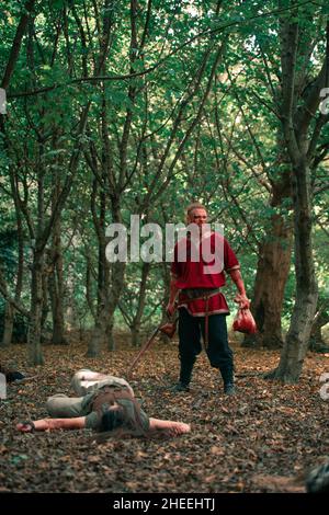 Corpo pieno di uomo aggressivo in costume vichingo con spada ucciso avversario steso a terra durante la rievocazione medievale Foto Stock