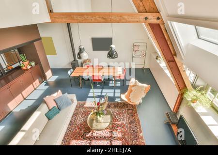 Dall'interno in alto di spazioso soggiorno contemporaneo con tavolo da pranzo circondato da sedie e tappeti sul pavimento e lampadari pendenti Foto Stock