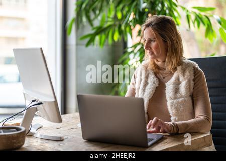 Concentrate le lavoratrici che digitano sul laptop mentre guardano il monitor del computer in un ambiente di lavoro moderno e leggero con piante verdi durante la giornata lavorativa Foto Stock