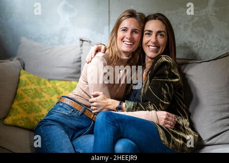 Le donne adorano i colleghi che abbracciano e guardano la fotocamera mentre si siedono su un comodo divano con cuscini in un moderno ufficio leggero durante la pausa Foto Stock