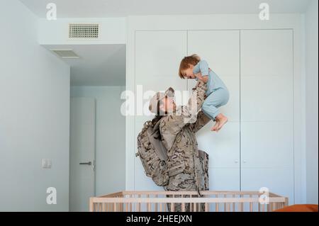 Vista laterale di felice soldato femminile in uniforme con zaino che tiene il figlio carino in braccia distese vicino al presepe in legno in camera da letto Foto Stock