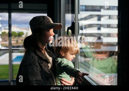 Vista laterale della soldato femminile in uniforme guardando fuori finestra con figlio carino mentre in piedi in sala luce con luce solare Foto Stock