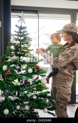 Felice soldato femminile in uniforme decorazione albero di Natale con figlio senza volto in soggiorno chiaro dopo essere arrivato dalla missione a casa Foto Stock