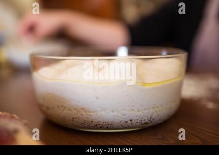 Pasta fresca bianca fatta in casa per la pizza in un recipiente di vetro posto su un tavolo di legno in una cucina leggera su sfondo sfocato Foto Stock