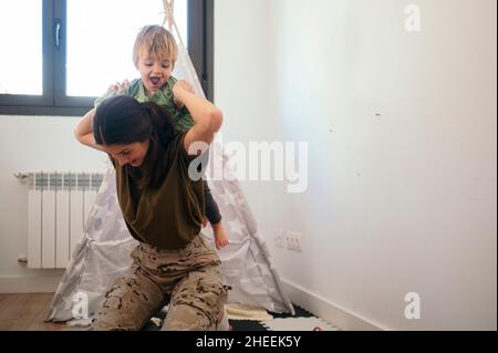 Donna militare positiva in uniforme che dà cavalcata piggyback al figlio carino mentre gioca insieme in sala giochi leggera con forte coperta Foto Stock