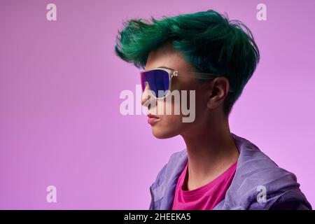 Vista laterale di elegante femmina con capelli corti tinti di verde in occhiali da sole con lente blu in piedi contro viola in studio Foto Stock