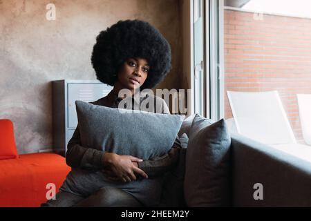 Donna afro-americana pensiva con acconciatura afro seduta su un comodo divano con cuscini mentre si guarda la finestra nella stanza luminosa Foto Stock