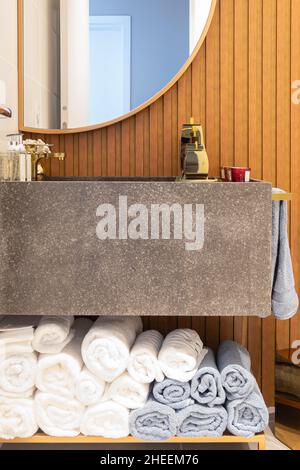 Un mucchio di asciugamani di cotone puliti posti su una mensola sotto il lavandino con prodotti da bagno e specchio appeso sulla parete di legno in bagno Foto Stock