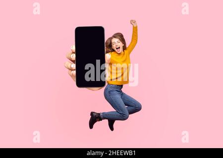 Felice giovane donna che vola e salta in aria e mostra grande schermo mobile vuoto per spazio copia e area pubblicitaria. Studio interno girato isolato su sfondo rosa Foto Stock