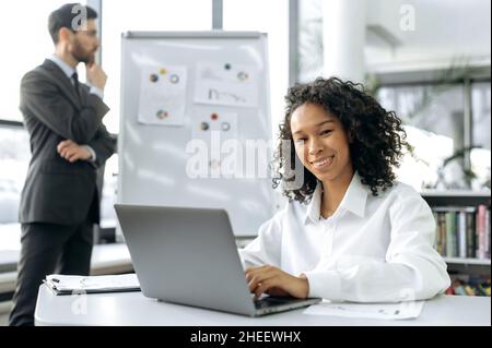 La giovane e intelligente donna d'affari afroamericana, manager, si siede a un tavolo in un ufficio moderno, usa un computer portatile, guarda nella fotocamera, sorride. In background un collega maschile studia grafici su un Foto Stock