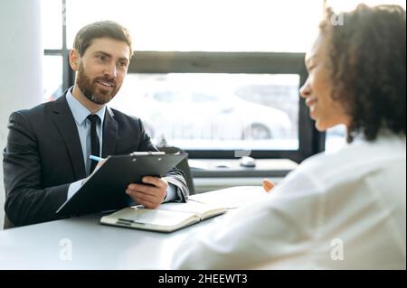 Intervista, concetto di assunzione. Caucasica successo CEO società, assumere manager, intervistando una donna afroamericana candidato, esaminando il suo curriculum, ascoltando le informazioni, in ufficio moderno, sorride Foto Stock