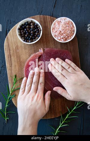 Preparazione del tonno con bistecca su sfondo di pietra nera. Mani femminili e un sacco di spezie, vista dall'alto. Foto Stock