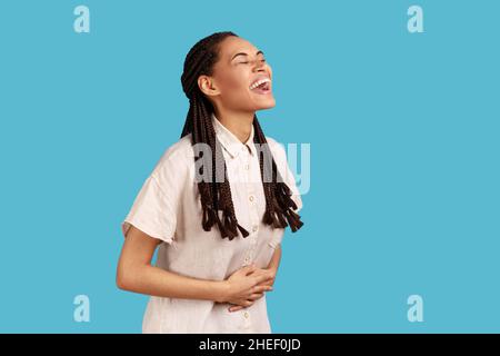 Ritratto di bella donna felice con dreadlocks neri che ride fuori forte, udendo scherzo divertente, esprimendo positivo, indossando camicia bianca. Studio interno girato isolato su sfondo blu. Foto Stock