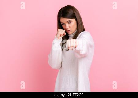 Donna aggressiva arrabbiata con capelli scuri pronti a combattere, boxe con fotocamera con pugni clenched, indossando un pullover casual bianco stile. Studio interno girato isolato su sfondo rosa. Foto Stock