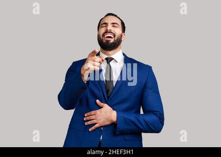Positivo eccitato uomo barbuto ridendo fuori forte tenendo il ventre e puntando il dito su di voi, beffe, indossando vestito di stile ufficiale. Studio interno girato isolato su sfondo grigio. Foto Stock