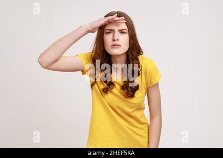 Curiosa ragazza adolescente con capelli marroni in T-shirt casual mantenendo la palma sopra la testa e guardando attentamente lontano, guarda con aspettativa a lunga distanza. Studio interno girato isolato su sfondo grigio. Foto Stock