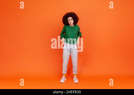 Ritratto a tutta lunghezza di donna con acconciatura Afro indossare maglione verde casual stile mostrando tasche vuote, non ha soldi, jobless femmina. Studio interno girato isolato su sfondo arancione. Foto Stock