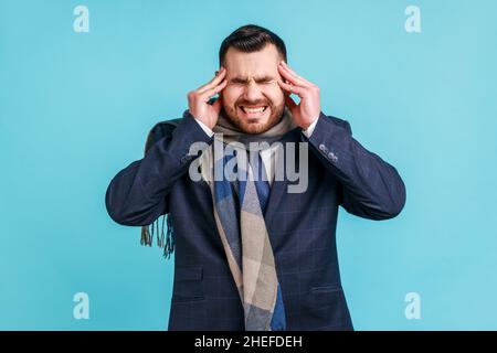 Ritratto di uomo malato bearded che indossa il vestito ufficiale di stile e avvolto in sciarpa che soffre di emicrania estremamente forte ed emicrania. Studio interno girato isolato su sfondo blu. Foto Stock