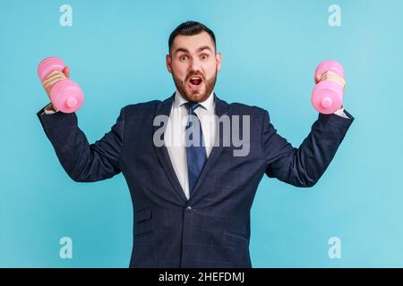 Forte uomo d'affari sicuro che indossa ufficiale abito da training bicipiti e triceps con manubri rosa, urlando, mostrando il suo potere. Studio interno girato isolato su sfondo blu. Foto Stock