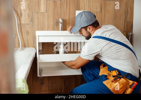 Idraulico professionale, lavoratore maschile in uniforme installazione lavello e tubo acqua in nuovo appartamento Foto Stock