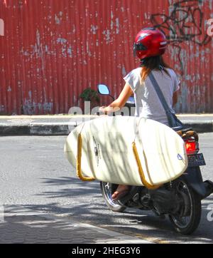 Una donna che guida una moto con una rastrelliera da surf che porta una tavola bianca a Kuta sull'isola di Bali in Indonesia. Foto Stock