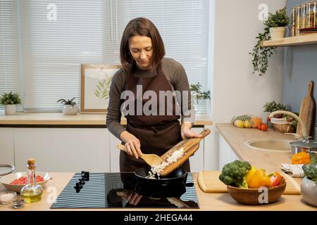 Giovane donna in grembiule che mette la cipolla fresca tritata in padella mentre cuoce la pasta italiana con un fornello elettrico Foto Stock