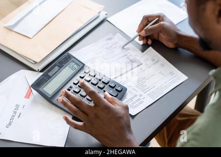 Mani di uomo africano premendo i pulsanti della calcolatrice mentre controlla i dati nei documenti finanziari e le fatture non pagate dalla tabella Foto Stock
