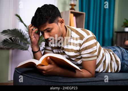 Confuso studioso college durante la lettura libro mentre dormendo sul divano - concetto di preparazione dell'ultimo giorno per l'esame, pigro studio e noioso studente. Foto Stock