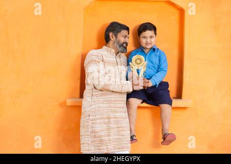 Ritratto giovane felice padre indiano con il suo figlio carino che tiene il premio vincente, uomo sorridente in vestito tradizionale in piedi con ragazzo in showin abito scuola Foto Stock