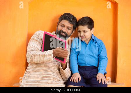 Felice padre indiano e il suo figlio piccolo carino in uniforme scuola scrittura su tavola di ardesia su sfondo arancione, adorabile bambino elementare con l'uomo barba Foto Stock