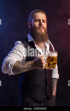 Ritratto di Bearded uomo che tiene la tazza di birra nella sua mano tatuaggi guardando la macchina fotografica su sfondo nero Foto Stock