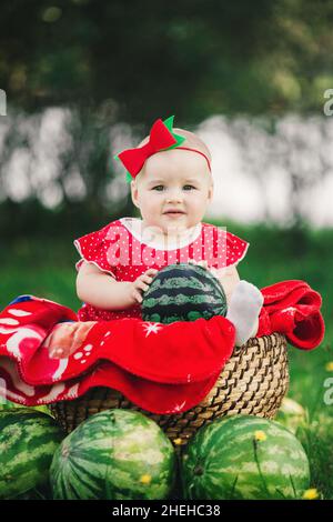 Carino 10 mesi bambina su pic-nic in erba con cocomeri. Sorridente bambino in abito rosso e arco sulla testa. Concetto di alimentazione sana, infanzia, frutta Foto Stock