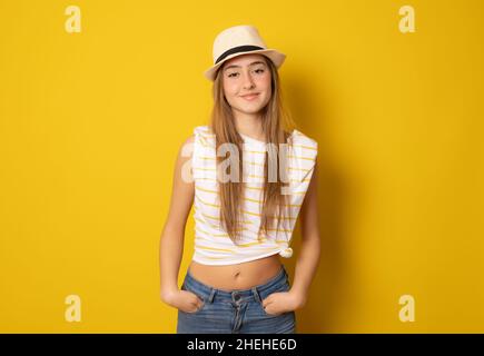 Giovane donna europea in piedi con le mani in tasche, con t-shirt casual a righe e cappello di paglia isolato su sfondo giallo. Foto Stock