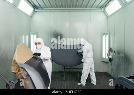 Giovane donna da lavoro in tuta e maschera protettiva in piedi vicino alla parte dell'auto, mentre il collega usa l'aerografo in garage Foto Stock