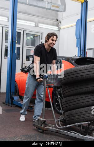 Meccanico allegro in piedi vicino auto con pneumatici e auto in garage Foto Stock