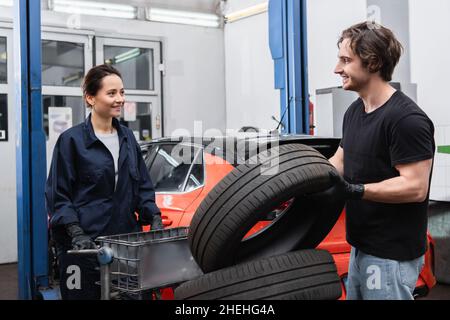 Meccanici sorridenti in piedi vicino agli pneumatici e auto sfocata in garage Foto Stock