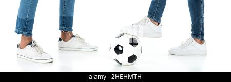 vista ritagliata di uomini interrazziali in jeans e sneakers vicino palla di calcio su bianco, banner Foto Stock