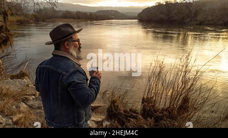 L'escursionista gode di uno splendido scenario naturale con una tazza di bevanda calda. Uomo che beve caffè e ammirare il bel fiume all'alba. Foto Stock