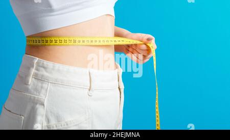 Donna sottile misura la sua linea vita con nastro di misurazione. Corpo sano che forma il concetto di perdita del peso. Vita sottile piccolo ventre in grandi pantaloni jeans bianchi Foto Stock