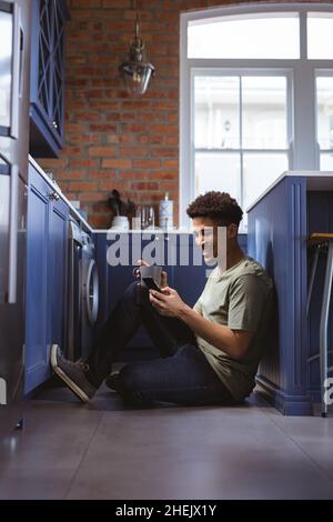 Sorridendo giovane biraciale che tiene la tazza di caffè con smartphone in cucina corridoio a casa Foto Stock