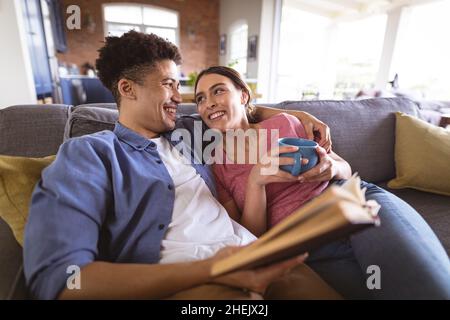 Felice coppia biraciale amorevole che si guarda l'un l'altro mentre si rilassa con caffè e libro a casa Foto Stock